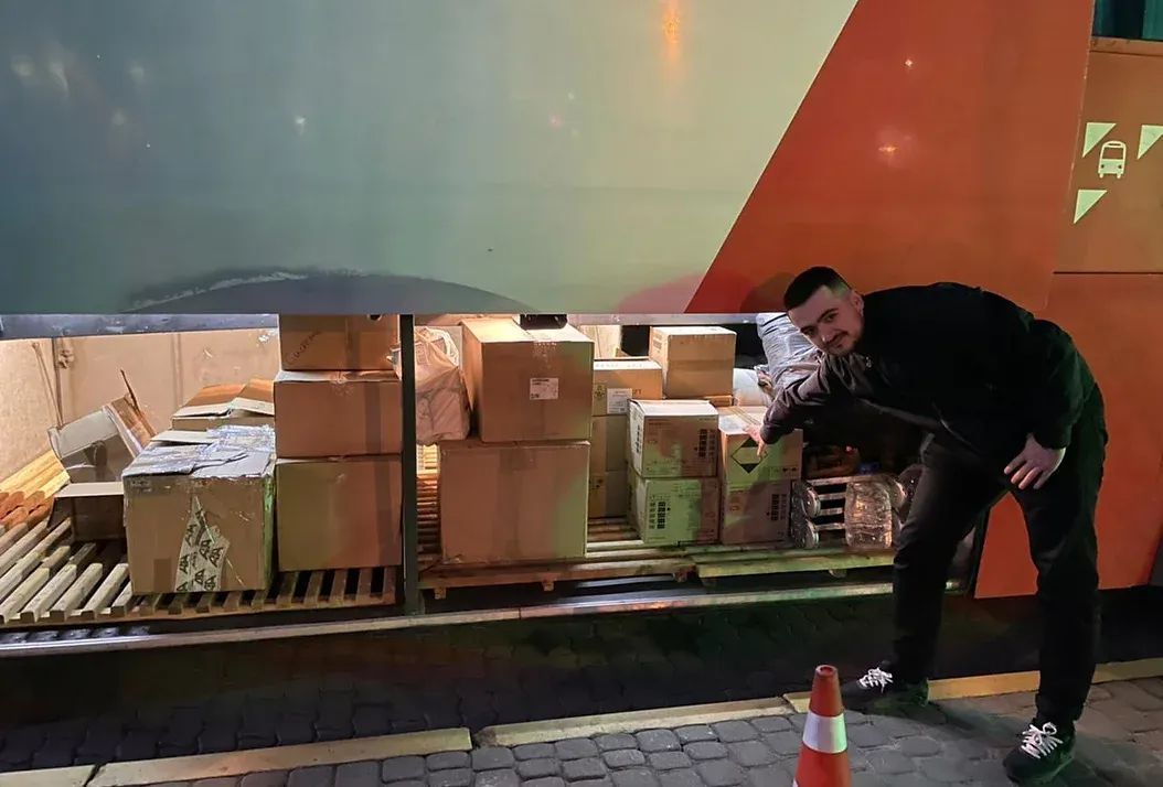 A man standing beside a bus full of humanitarian aid items in cardboard boxes.