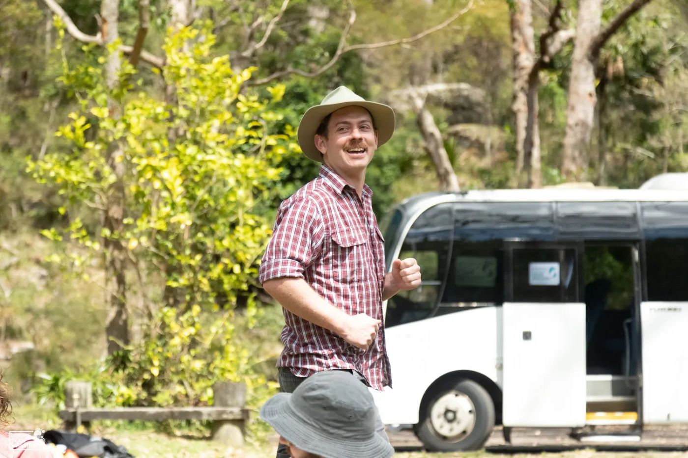 Man in a flannel shirt wearing a hat in front of a bus, smiling